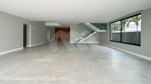 Sala grande em tres ambientes com parede e piso cinza, escadas com proteção de vidro, à venda da barra da tijuca