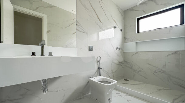 banheiro da primeira suíte, com porcelanato branco, espelho e um chuveiro, à venda na barra