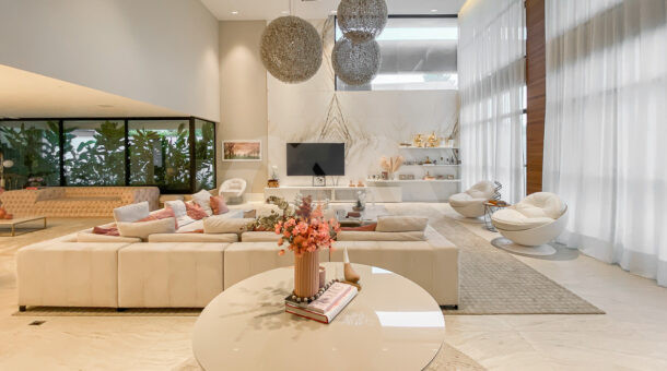 Sala ampla de casa à venda com pé direito alto, com extenso sofá, decoração clean com tons de rosa e grandioso lustre