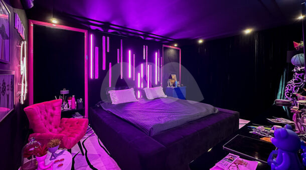 Quarto com paredes pretas, luz neon em tom roxo e rosa, cama acolchoada, poltrona e painel para TV