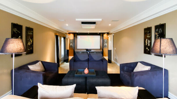 Sala de cinema com projetor, sofás e poltronas em tom de azul e bege em casa à venda na Muller Imóveis RJ