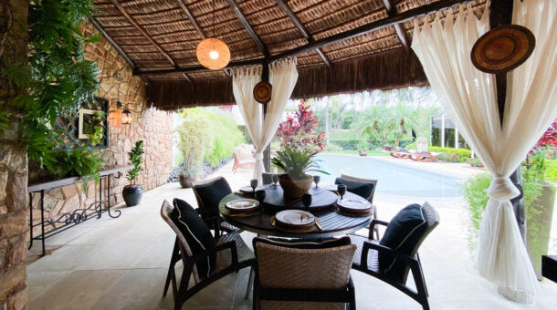 área gourmet de casa à venda no Malibu com teto em palha, mesa de madeira redonda e cortinas para mais privacidade
