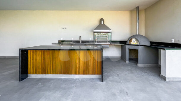 Área gourmet com bancada com mármore preto e detalhes de madeira, com uma churrasqueira e forno de pizza, à venda no alphaville