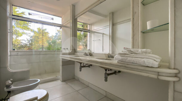 Imagem de banheiro com bancada dupla branca e chuvei com ampla janela com vista para jardim da casa triplex a venda na Muller Imoveis RJ