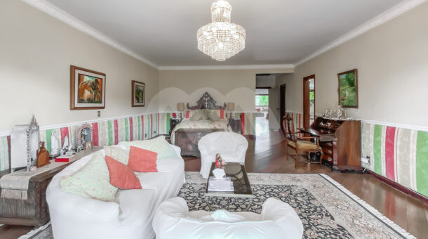 Imagem de quarto com papel de parede listrada, sofá branco com lustre e cama de casal em casa a venda na Barra da Tijuca