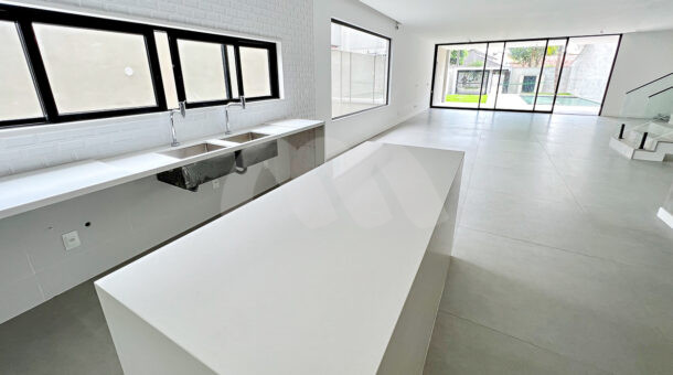 Imagem de cozinha com ilha branca e bancada com pia dupla em casa à venda na Barra da Tijuca
