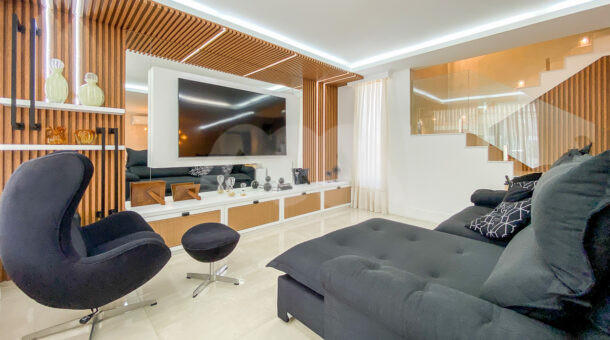 Imagem de sala ampla com painel de tv em madeira ripada com projeto de iluminação, e poltrona com pufe